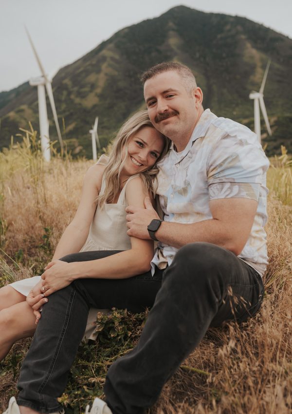 Couple embracing during a candid engagement photoshoot with mountains and wind turbines in the background.
